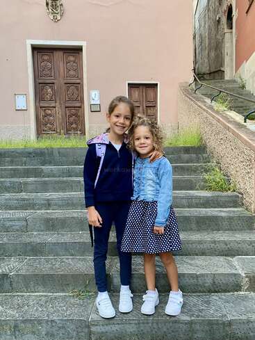 Deux jeunes filles se tiennent sur des marches en pierre, souriantes, les bras enlacés. Elles portent des tenues décontractées et des baskets blanches, et posent devant un vieux bâtiment.