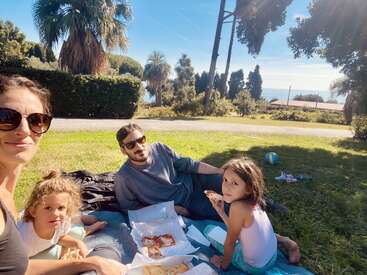 Une famille de quatre personnes profite d'un pique-nique ensoleillé sur une couverture, en mangeant une pizza en plein air. Les palmiers, le ciel bleu et la mer au loin créent une atmosphère détendue et joyeuse.