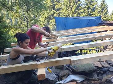 Two people are working together on constructing a wooden frame outdoors. Surrounded by trees, they use tools and wear gloves, focused on building a sturdy structure.
