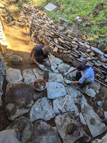 Two people are carefully laying large stone slabs to create a pathway, surrounded by a dry-stone retaining wall, buckets, and construction tools in an outdoor setting.