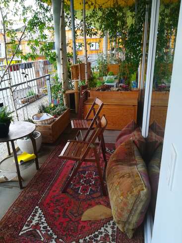 This cozy balcony features wooden chairs, a red patterned rug, large cushions, potted plants, trees, and wooden planters, creating a lush and inviting outdoor retreat.