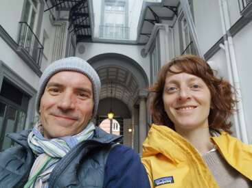 A smiling man and woman take a selfie in a beautiful arched corridor. He wears a beanie and scarf; she wears a yellow jacket. Bright, cheerful atmosphere.
