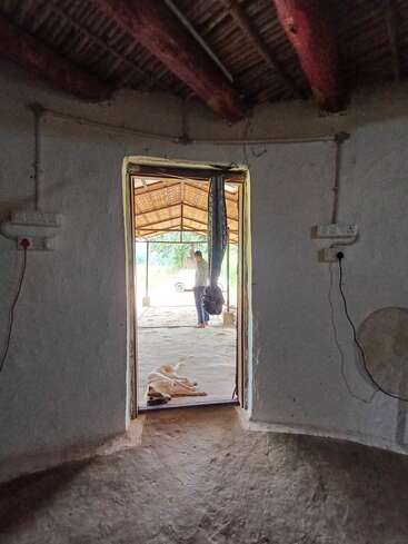 Esta imagem mostra um cômodo rústico, com piso de barro e telhado de palha. Um cachorro está na porta, do lado de fora uma pessoa está sob um dossel de madeira.