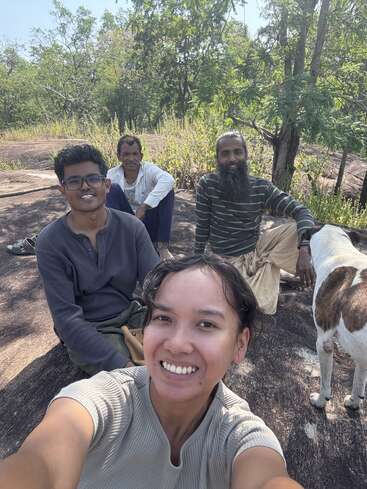 Quatro pessoas e um cachorro sentados ao ar livre em uma superfície rochosa, cercados por árvores e vegetação, sorrindo e desfrutando da companhia uns dos outros em um dia ensolarado.