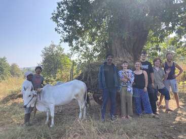 La imagen muestra a un grupo de siete personas de pie frente a un árbol, con una vaca blanca a la izquierda, sobre un fondo de árboles y cielo despejado.