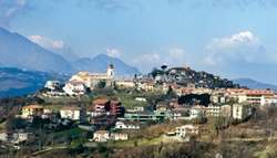 The image depicts a small town situated on a hill, with a church tower visible in the distance, set against a backdrop of mountains and a blue sky.