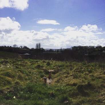 A imagem mostra um campo gramado com uma pequena ponte de madeira e dois canos vermelhos em primeiro plano, em um céu azul com nuvens brancas.