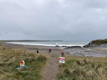 A cloudy beach scene with grassy dunes and rocks, a few people scattered, waves crashing, and a "No Swimming" warning sign prominently displayed near the shore.