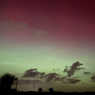 Un ciel nocturne serein, baigné de reflets verts et violets, sans doute dus à une aurore boréale, surplombe un paysage rural où se dessinent les silhouettes d'arbres, de lignes électriques et de nuages sombres.