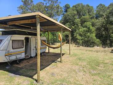 Uma caravana branca está estacionada sob um abrigo de madeira com uma rede pendurada ao lado. O cenário é gramado, cercado por árvores altas e verdes e céu azul.