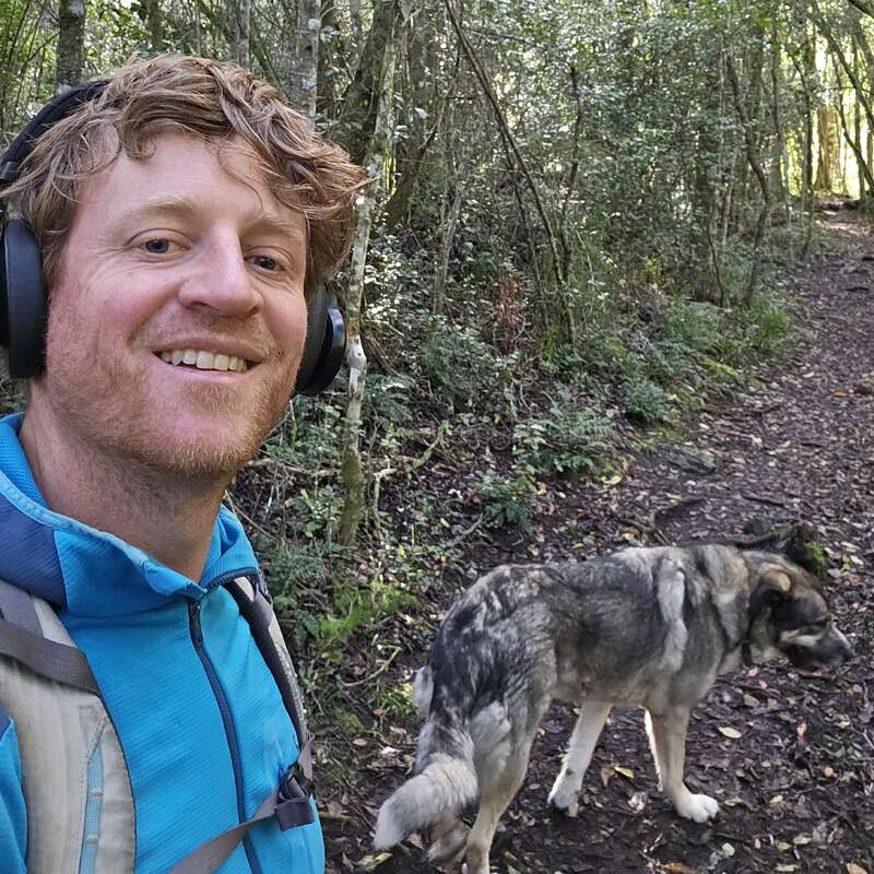 Um homem usando fones de ouvido e uma jaqueta azul sorri para uma selfie enquanto caminha em uma área de floresta com seu cachorro parecido com um lobo caminhando ao seu lado.