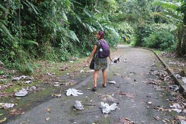 A woman with a purple backpack walks along a leaf-strewn, deserted road surrounded by lush green jungle, creating a sense of adventure and exploration.