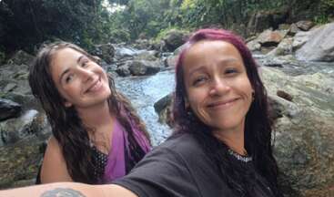 Two women smile happily while taking a selfie by a rocky river in a lush, green forest. Both appear relaxed, enjoying nature and each other’s company.
