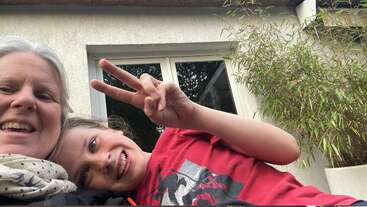 Eine lächelnde Frau und ein Kind machen ein Selfie im Freien. Das Kind, das ein rotes T-Shirt trägt, zeigt ein Friedenszeichen. Hinter ihnen stehen Grünpflanzen und ein Fenster.