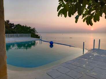 A tranquil infinity pool overlooks a calm sea at sunset, with reflections shimmering on the water, leafy branches above, and a serene, peaceful atmosphere throughout.