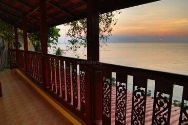 A beautiful wooden balcony overlooks a tranquil sea at sunset. Ornate railings, lush trees, and serene water create a peaceful, tropical atmosphere perfect for relaxation.