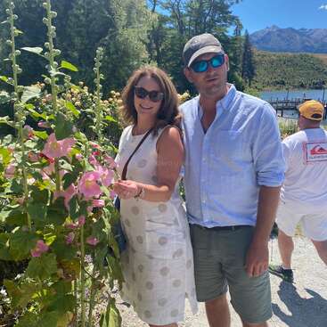 Una pareja sonriente posa junto a altas flores rosas en un día soleado. Detrás de ellos hay árboles, un lago, montañas y una persona con sombrero amarillo.