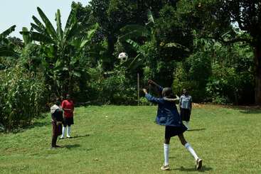 Quatro crianças estão jogando futebol em um campo gramado cercado por árvores verdes exuberantes e bananeiras, com uma criança cabeceando ativamente a bola no ar.