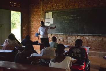 Um professor está na frente de uma sala de aula rústica, instruindo alunos atentos sentados em carteiras de madeira. As lições e anotações estão escritas em um grande quadro-negro.