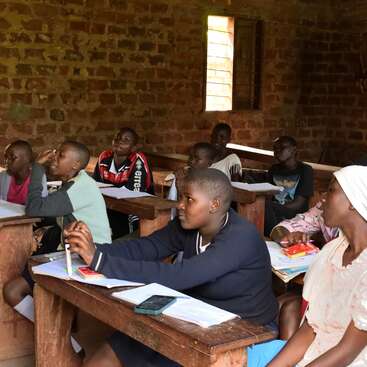 Un grupo de estudiantes atentos, sentados en pupitres de madera en un aula con paredes de ladrillo y una ventana abierta, se dedican a aprender y a tomar apuntes.
