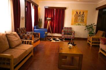 Esta aconchegante sala de estar apresenta poltronas coloridas, uma mesa de centro de madeira, cortinas marrons, iluminação aconchegante, plantas, obras de arte africanas e um tapete estampado em um piso de ladrilhos.