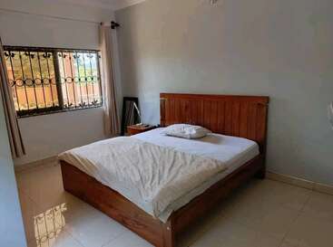 Um quarto simples com cama de madeira, roupa de cama branca, travesseiro único, mesa de cabeceira, espelho, janela com grades e cortinas e piso de ladrilho limpo e de cor clara. Decoração minimalista.