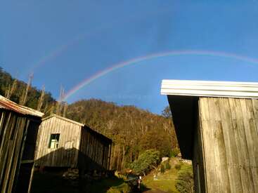 The image depicts a serene scene of a rainbow arcing over a rustic wooden building, set against a backdrop of trees and a clear blue sky.