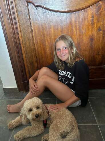 A young girl with blonde hair sits on a tiled floor, smiling beside a fluffy, light brown dog. They are indoors, against a wooden door, looking content.