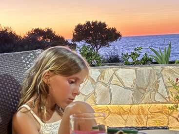 A young girl sits pensively at an outdoor table. Behind her, a beautiful stone wall, lush plants, and a vibrant sunset over the ocean complete the scene.