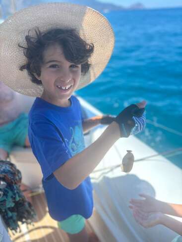 A smiling child with curly hair and a wide-brimmed straw hat stands on a boat, holding a small fish, enjoying a sunny day on the bright blue ocean.