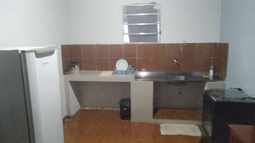 This image shows a simple kitchen with a refrigerator, sink, stove, brown tiled backsplash, dish rack, trash bin, window, and a small shelf above the sink.