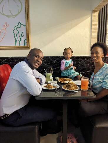 A smiling family of three sits together at a restaurant table, enjoying a meal. The parents are happy, and their young daughter looks curiously at the camera.