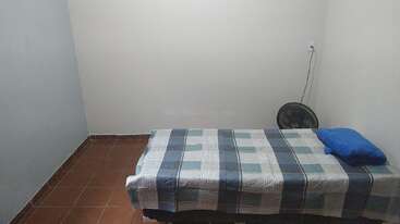 This image shows a simple, small room with a single bed covered by a checkered blanket, a blue pillow, a fan, and plain tiled floor.
