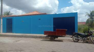 A bright blue building with a tiled roof stands beside a paved street. A black motorcycle with a red trailer attachment is parked near the sidewalk. Palm trees, cloudy sky.