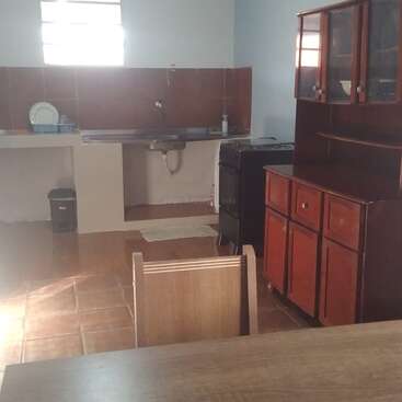 This image shows a simple kitchen with a wooden cabinet, a table, a stove, a sink area, a dish rack, brown tiles, and natural light.