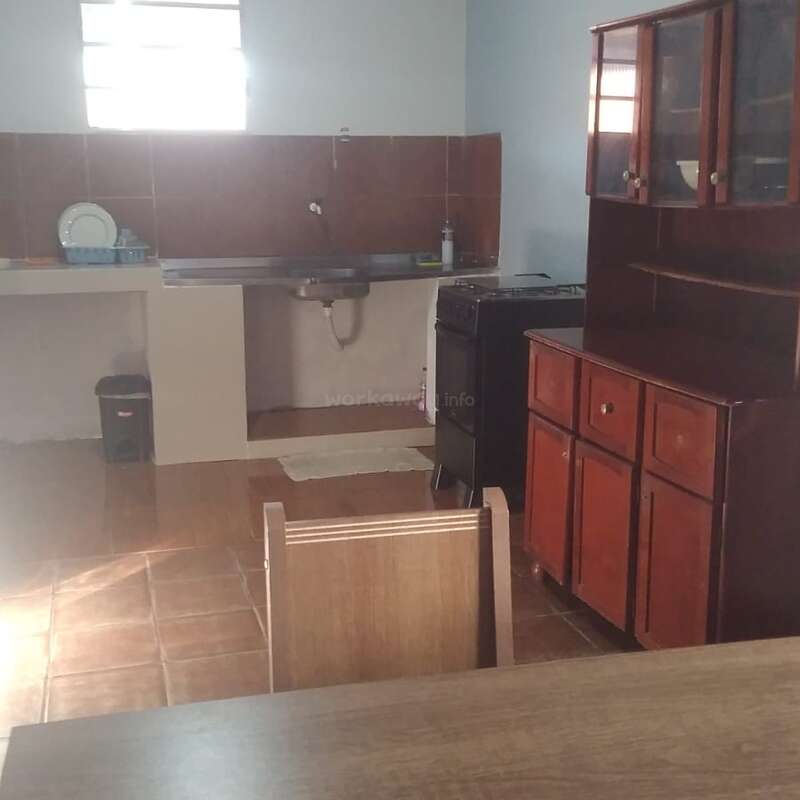 This image shows a simple kitchen with a wooden cabinet, a table, a stove, a sink area, a dish rack, brown tiles, and natural light.