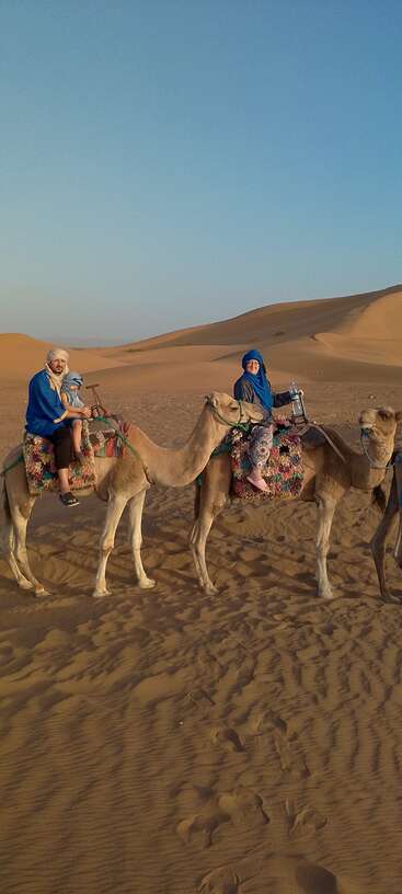 Three people dressed in blue ride camels across vast desert sand dunes under a clear blue sky, creating a serene and adventurous scene in the golden evening light.