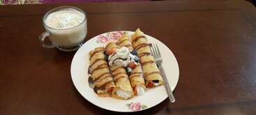 Three rolled crepes topped with chocolate drizzle, whipped cream, blueberries, and strawberries are served on a floral plate, accompanied by a cup of frothy coffee.