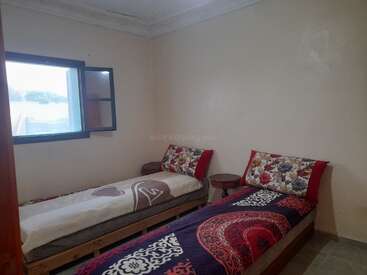 This image shows a simple bedroom with two single beds, colorful bedding, floral pillows, a small wooden nightstand between them, beige walls, and an open window.