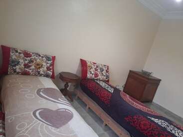 This image shows a simple bedroom with two single beds, floral pillows, colorful blankets, a small round wooden table, and a wooden dresser in the corner.