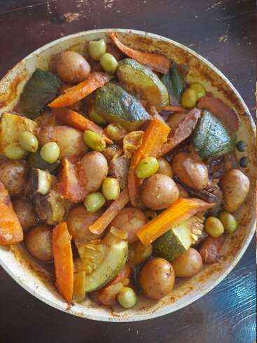 A hearty stew featuring baby potatoes, sliced carrots, zucchini, green olives, and chunks of meat in a flavorful, richly colored broth, served in a round dish.