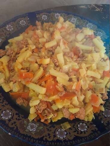 On a decorative blue plate, a colorful vegetable and meat stir-fry features chopped potatoes, carrots, cabbage, tomatoes, and ground meat. Steam rises, indicating it's freshly cooked.
