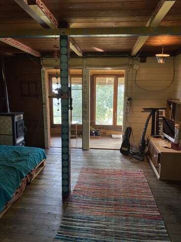 This cozy wooden room features a bed, a colorful rug, guitar, TV, wood stove, painted beams, large windows, and eclectic, artistic, and homely decorations throughout.
