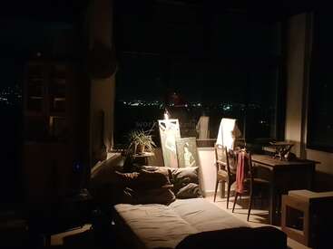 A cozy, dimly-lit room with a sofa bed, desk, two chairs, a plant, and artwork. Large windows reveal city lights glowing in the night.