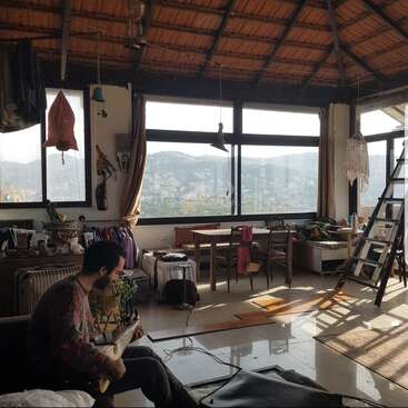 A man plays guitar in a cozy, sunlit room with large windows, wooden ceiling, eclectic decor, plants, and a scenic mountain view in the background.