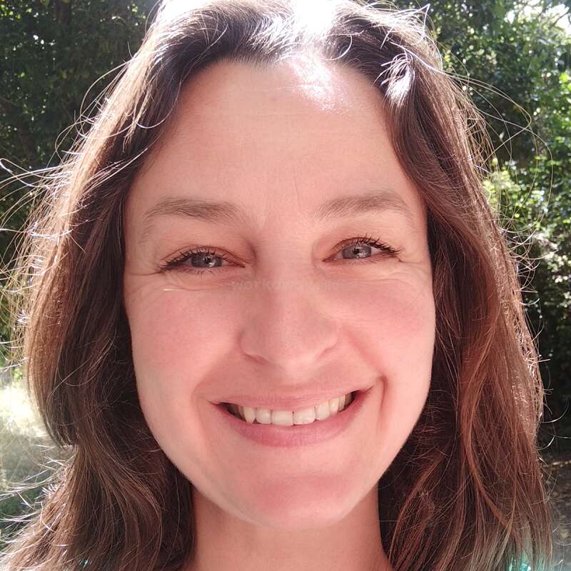 A woman smiles warmly outdoors, sunlight illuminating her hair. Green trees and bright light create a cheerful, natural background. Her happy expression radiates positivity and friendliness.