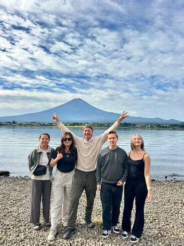 Cinco amigos posan en la orilla rocosa de un lago, con el monte Fuji al fondo, sonriendo bajo un cielo nublado y lleno de color, mientras disfrutan juntos de un precioso momento al aire libre.