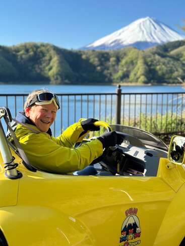 Una persona sonriente, vestida con una chaqueta amarilla y gafas protectoras, conduce un descapotable amarillo junto a un lago, con el monte Fuji nevado y unas exuberantes colinas al fondo.