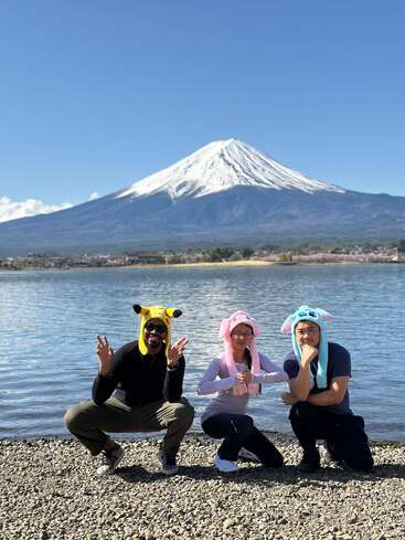 Tres personas con divertidos sombreros de animales posan alegres a orillas de un lago, con el majestuoso monte Fuji, cubierto de nieve, elevándose imponente sobre el fondo de un cielo azul claro.