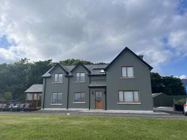 This image shows a large, two-story grey house with a wooden door, multiple windows, a spacious lawn, outdoor seating, trees, and a cloudy sky.